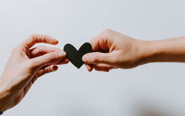 Two hands hold either side of a small, black paper cut out heart.